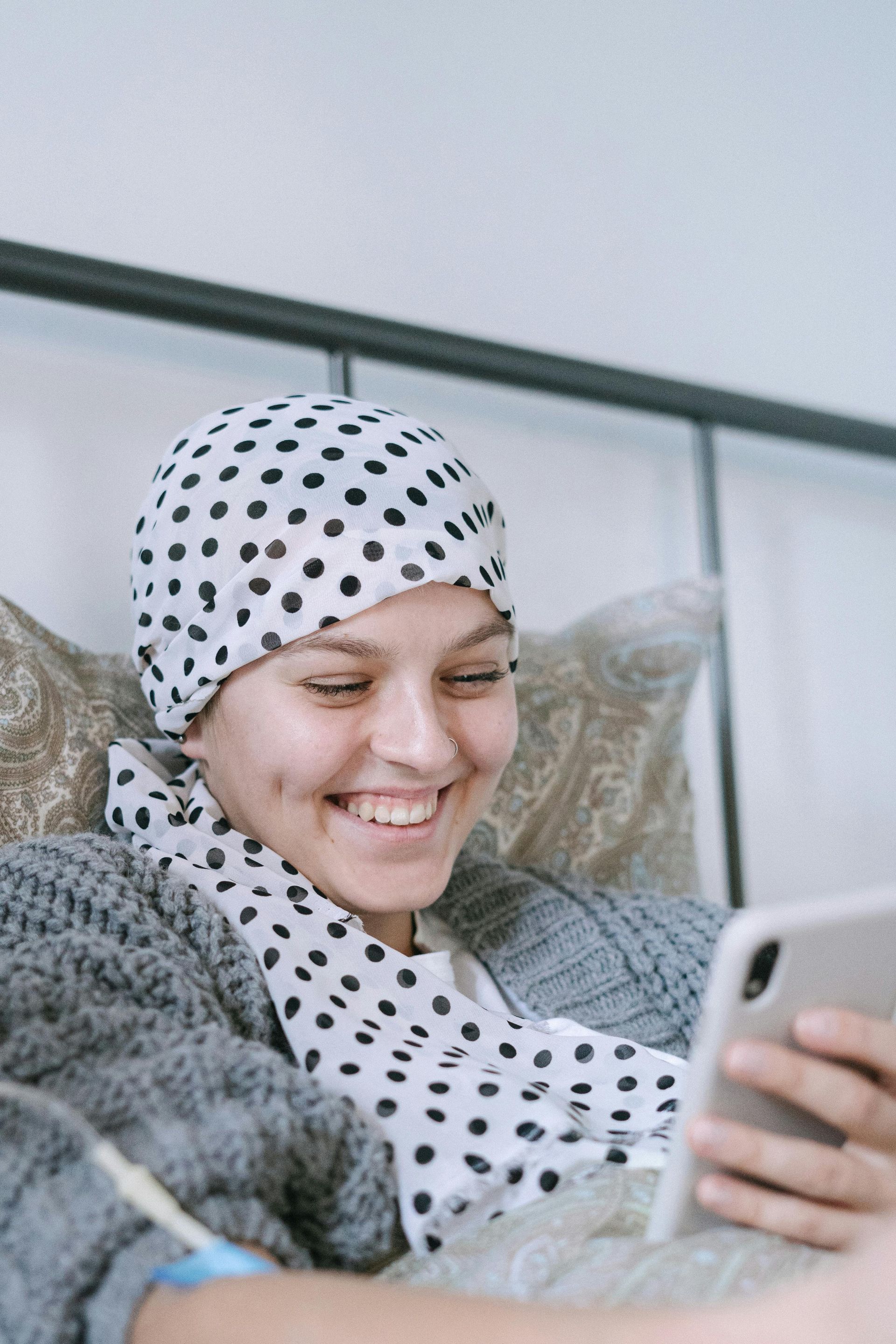 Woman with a polka-dot headscarf smiles while looking at her phone in a bed.