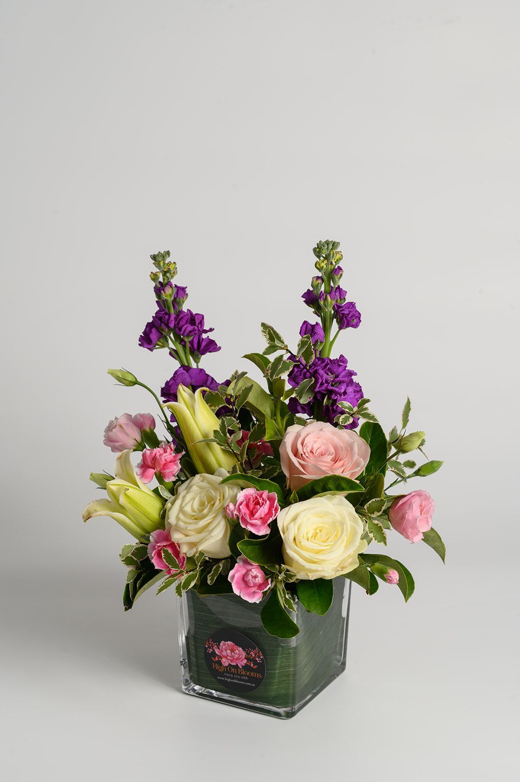Floral arrangement in a clear square vase: white and pink roses, purple stock flowers, and green foliage.
