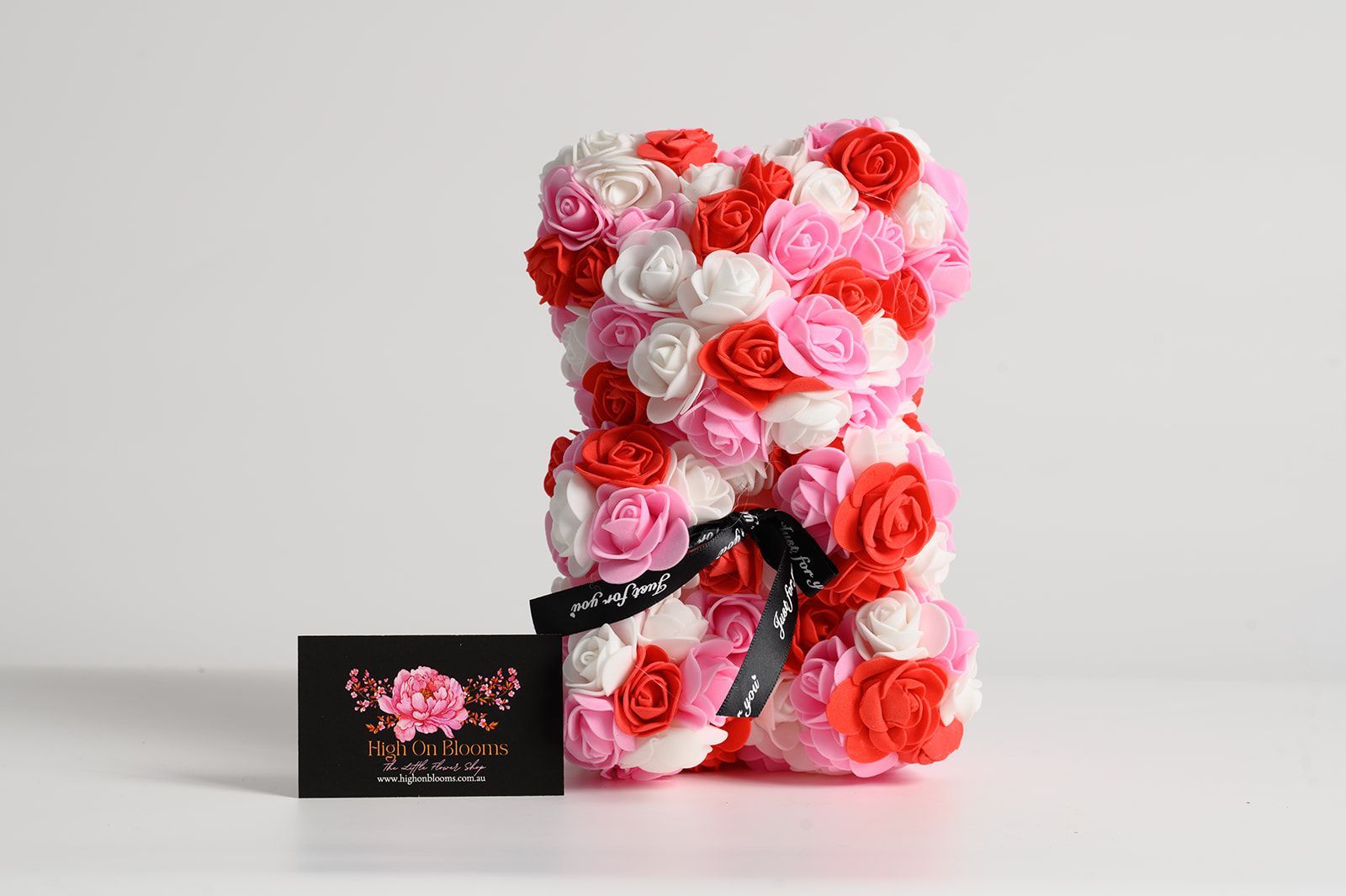 Teddy bear sculpture made of pink, white, and red roses, with a black ribbon. A black card sits in front.