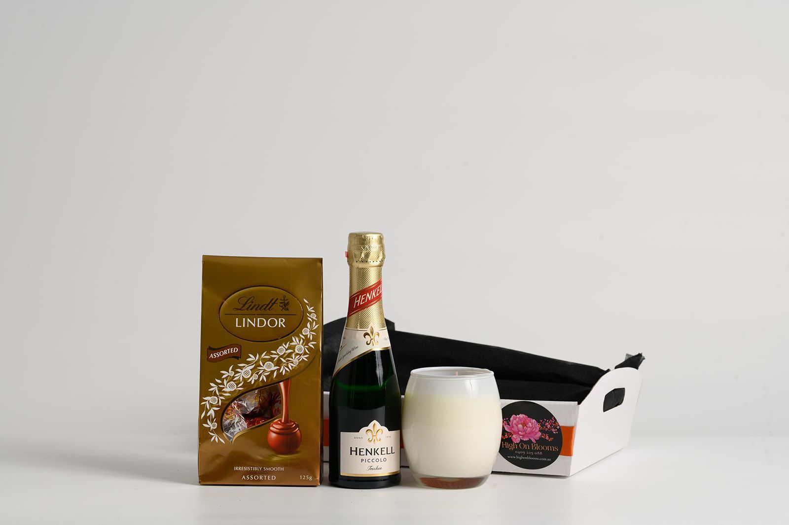 Gold-wrapped chocolate box, champagne bottle, frosted glass, and gift box in a white tray against a white background.