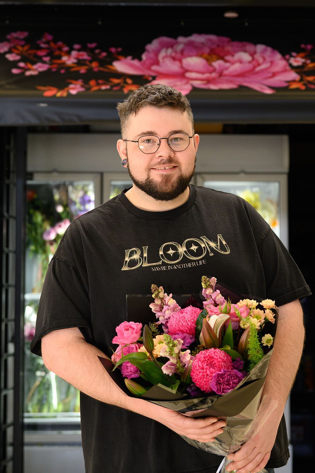 Man holding bouquet, smiling, wearing glasses and black t-shirt. Flowers and floral shop background.