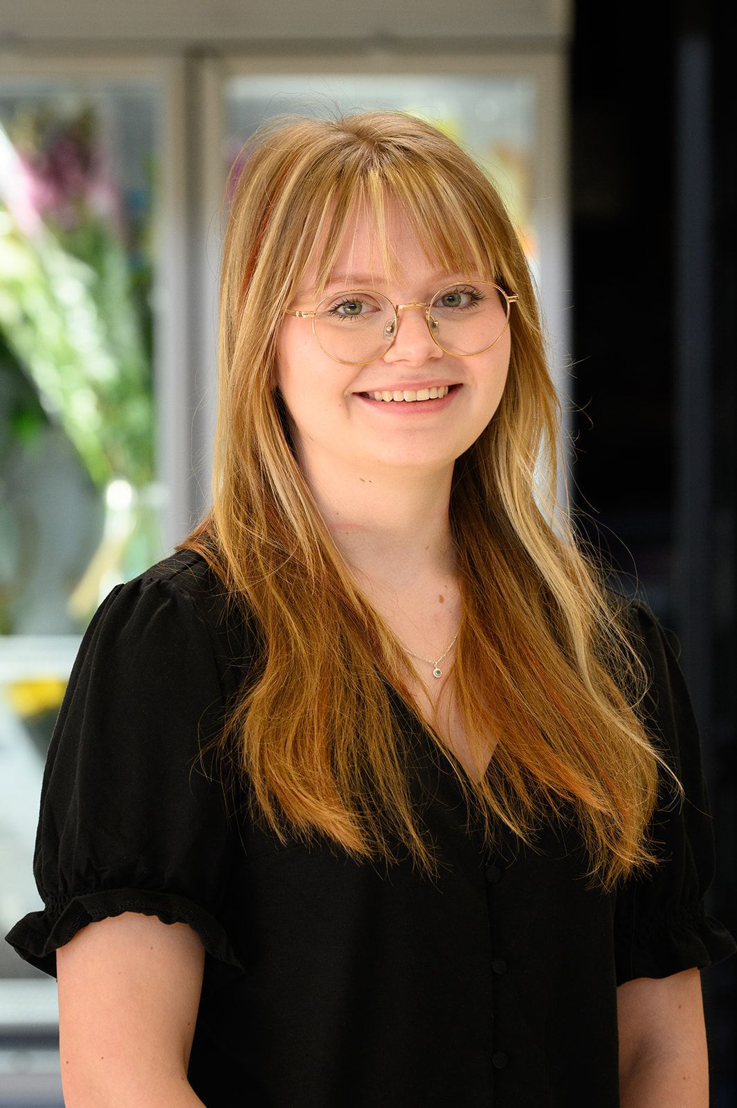 Woman with long blonde hair, glasses, and a black shirt smiling in front of a blurred background.