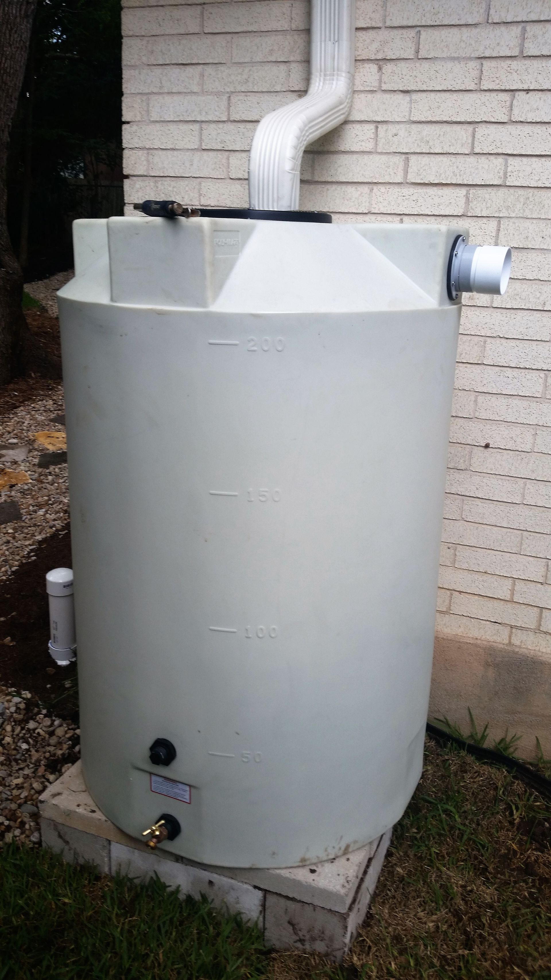A gray rainwater harvesting tank with a white downspout connected to it, placed against a brick wall.
