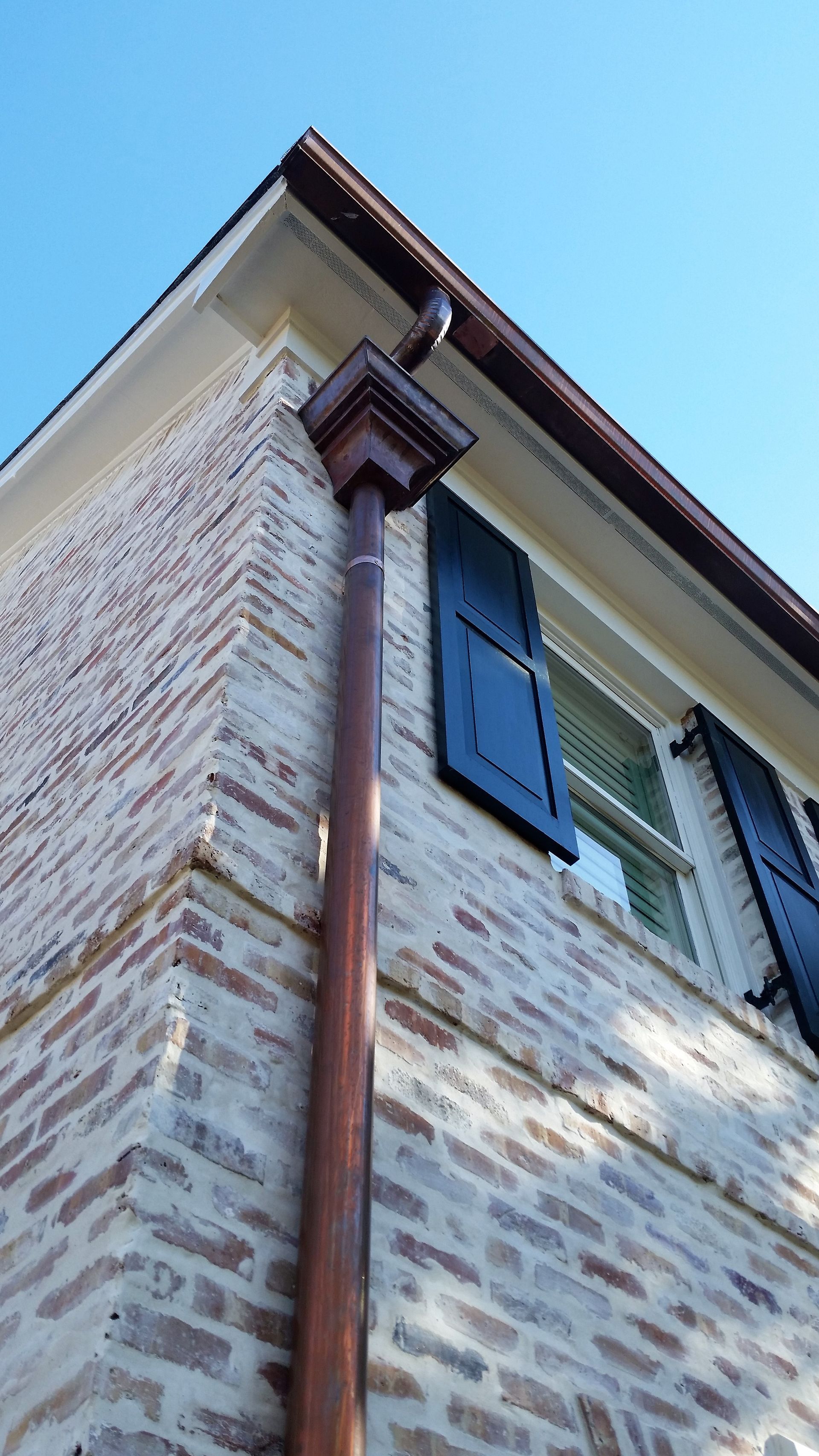 Corner of a brick building with copper gutters and dark shutters against a blue sky.