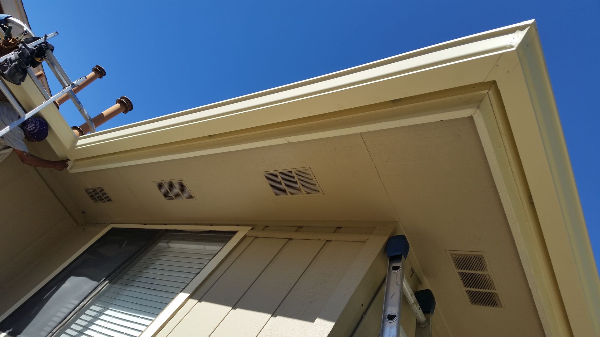 Beige house exterior with gutters and vents against a blue sky, ladder visible.