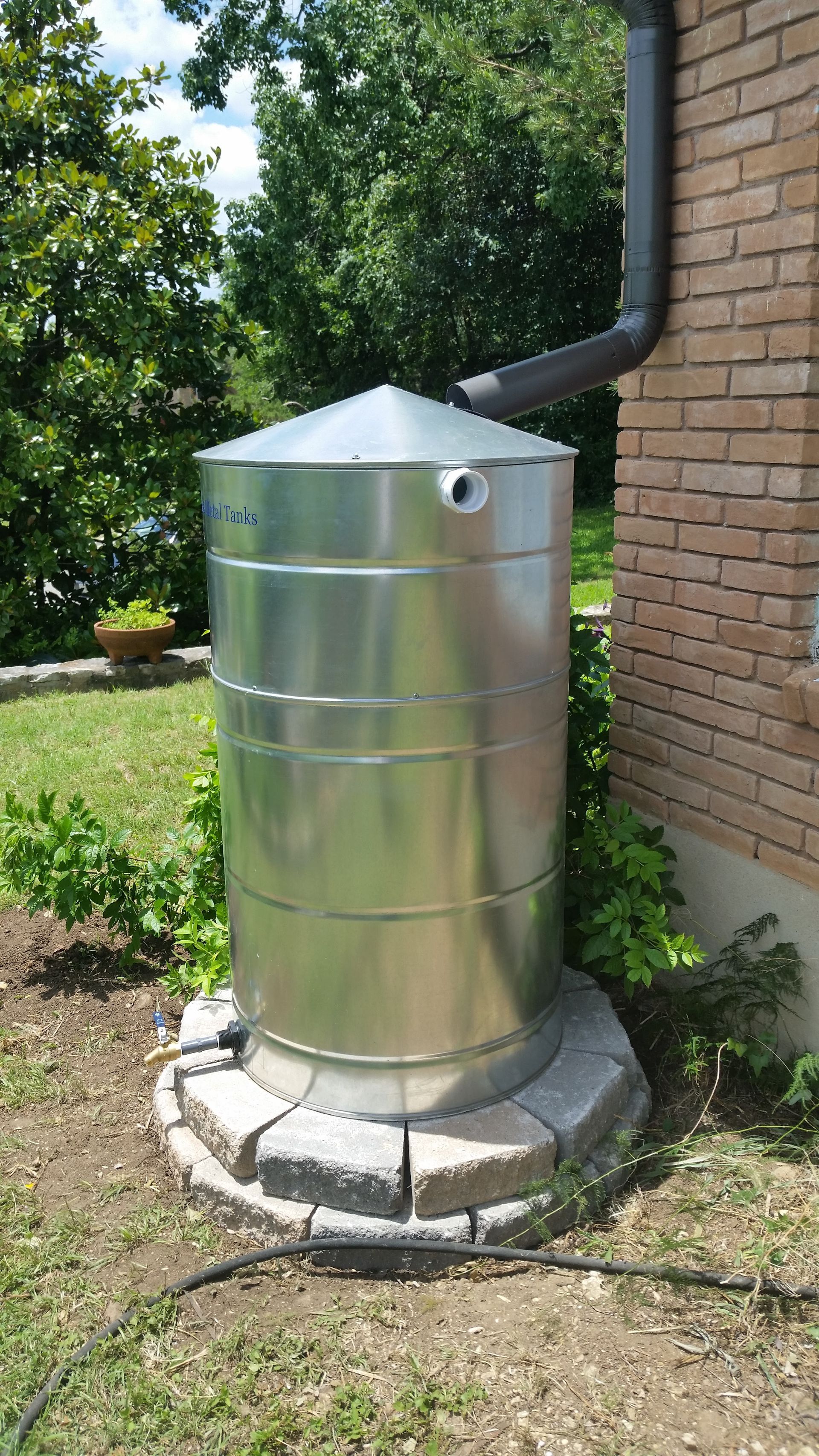 Metal rainwater collection tank next to a brick building, with a downspout and stone base.