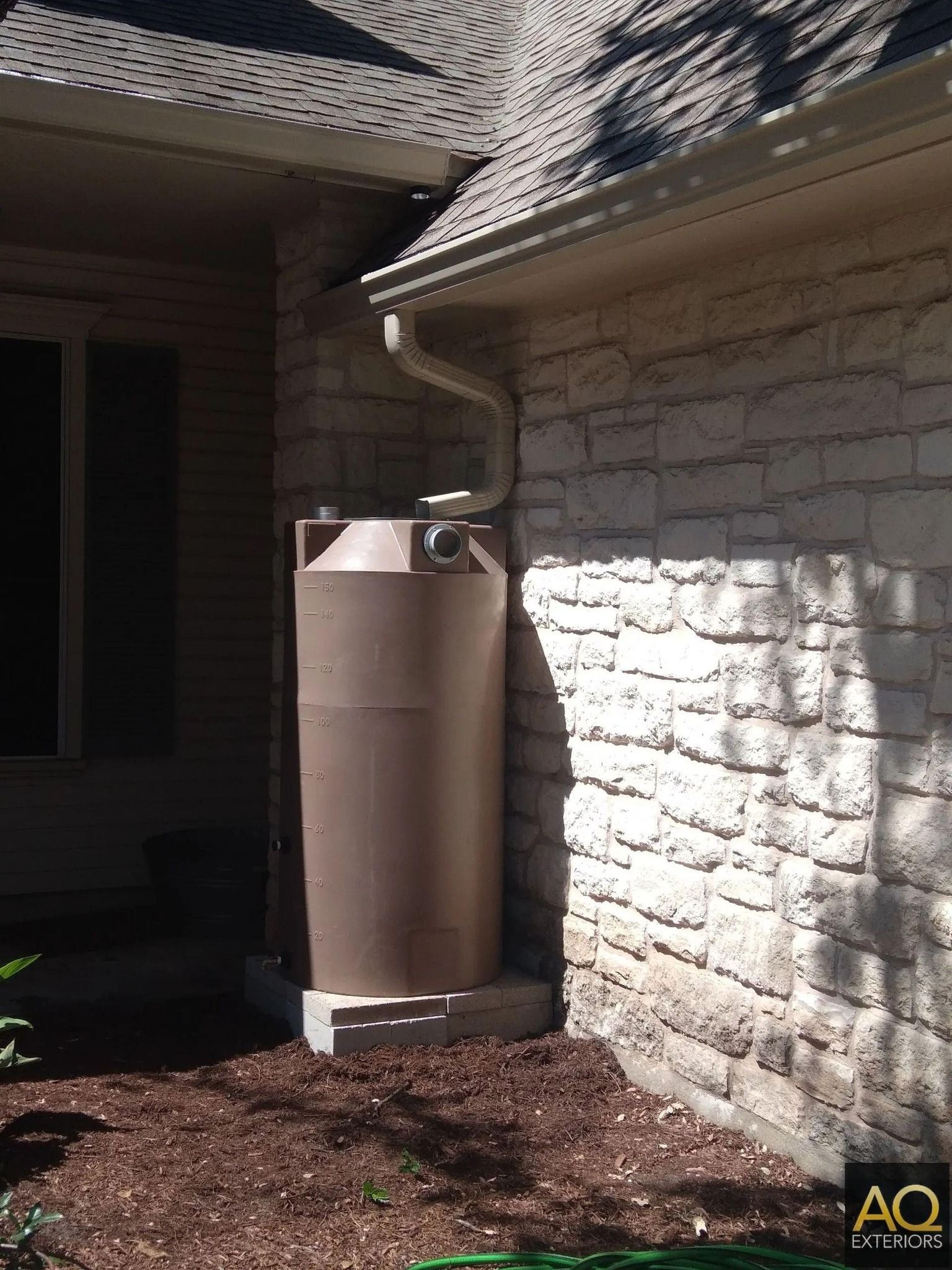 Brown rain barrel next to a beige brick house, connected to a gutter, set on a concrete base.
