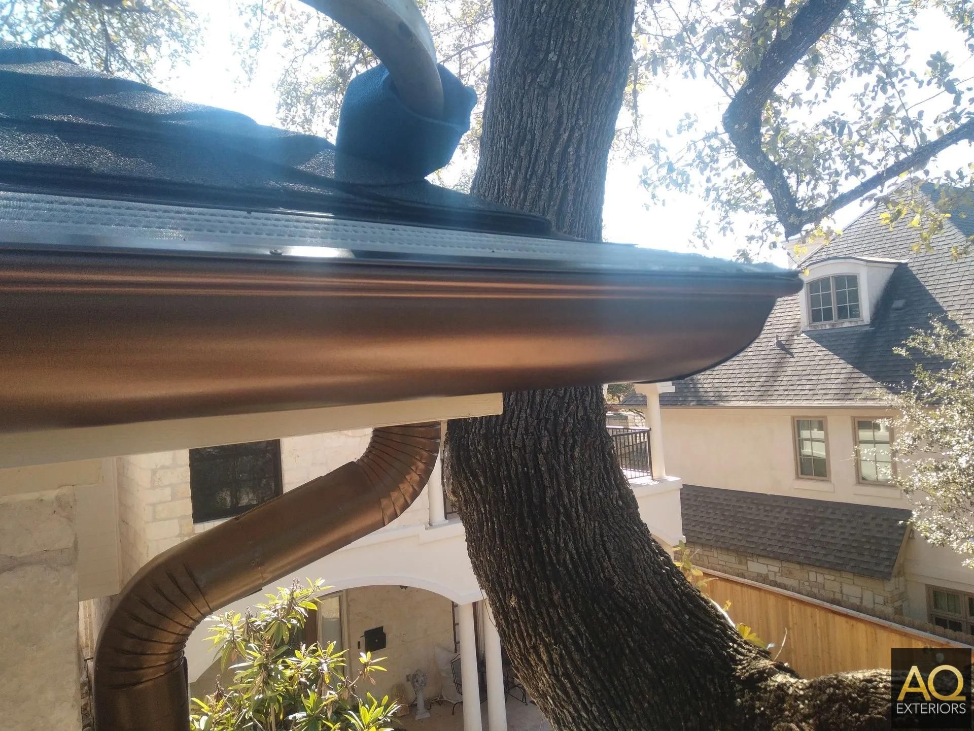 Bronze gutters on a house, a tree trunk partially blocking the view. The sky is bright.