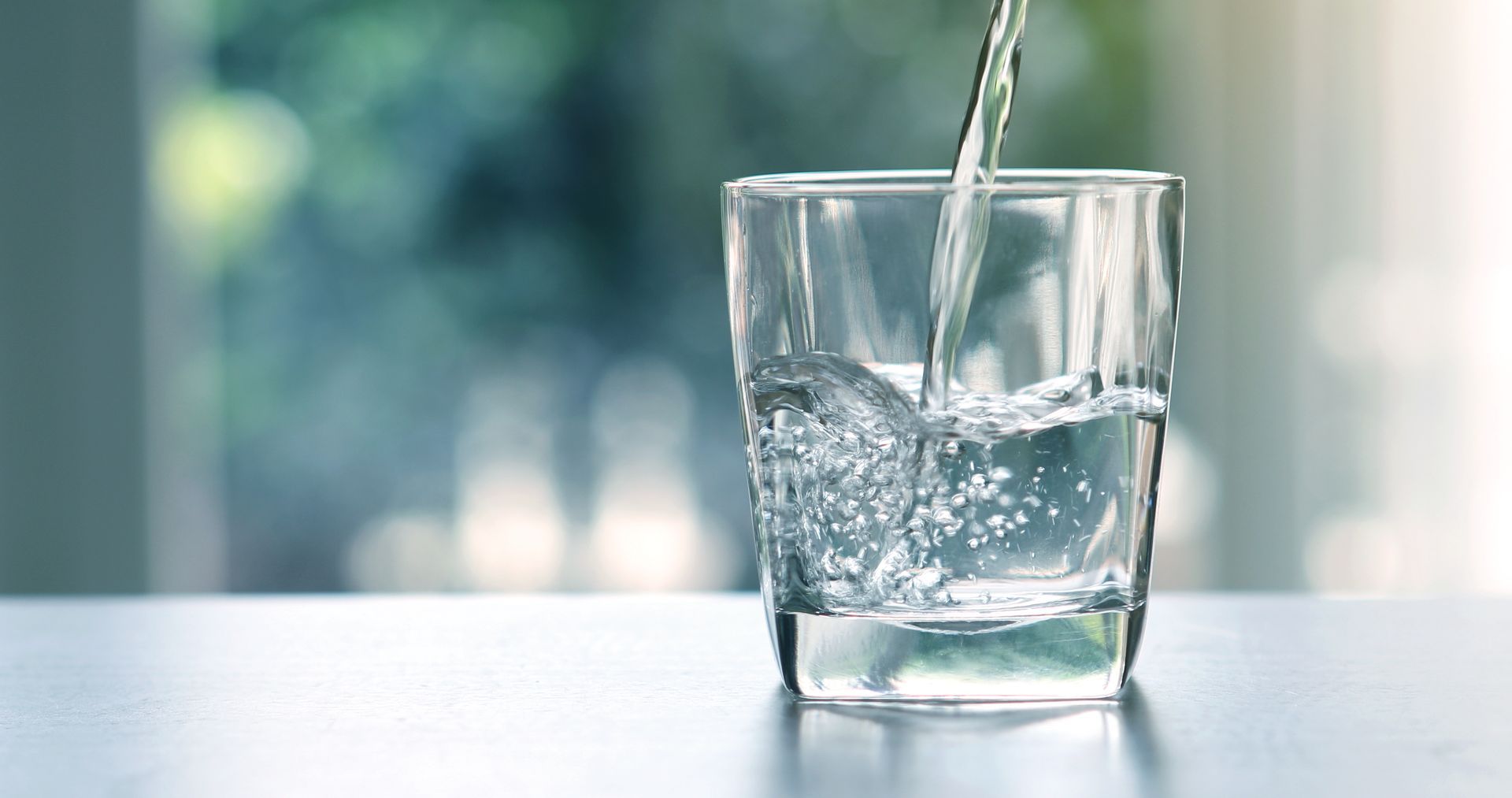 Clear water pours into a transparent glass sitting on a table against a soft, blurred background.