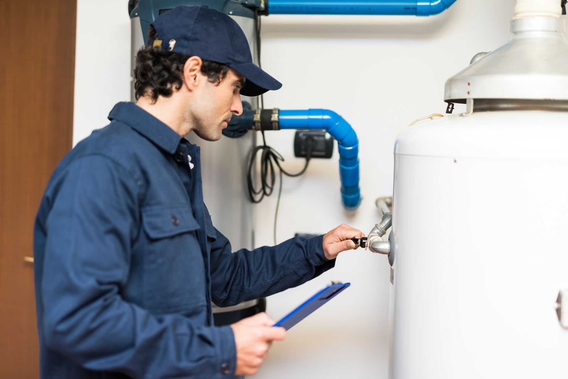 A person in a blue uniform and cap inspects a white water heater while holding a clipboard.
