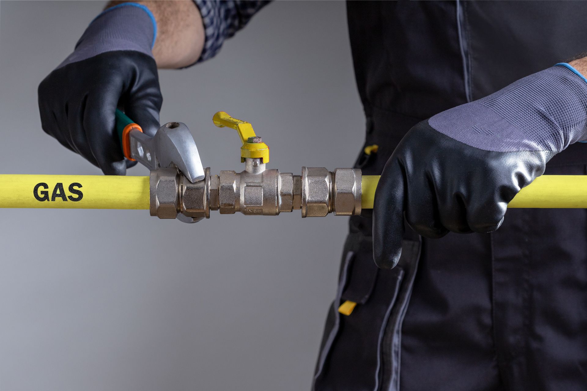 A person wearing work gloves uses an adjustable wrench to tighten a metal valve on a yellow pipe labeled 