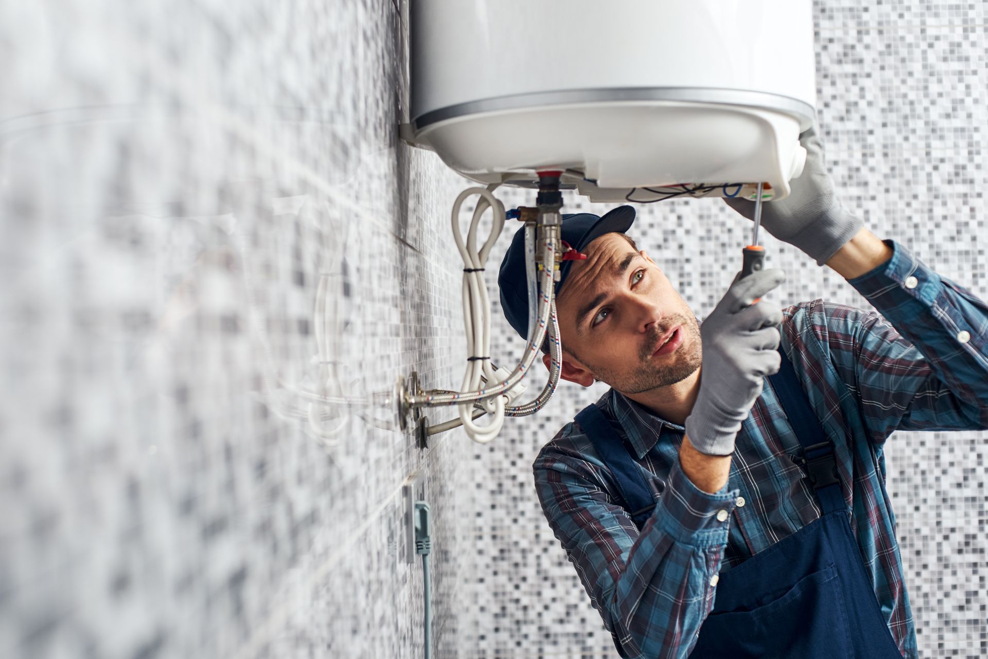 A person in work clothes and gloves using a screwdriver to repair a white wall-mounted water heater.
