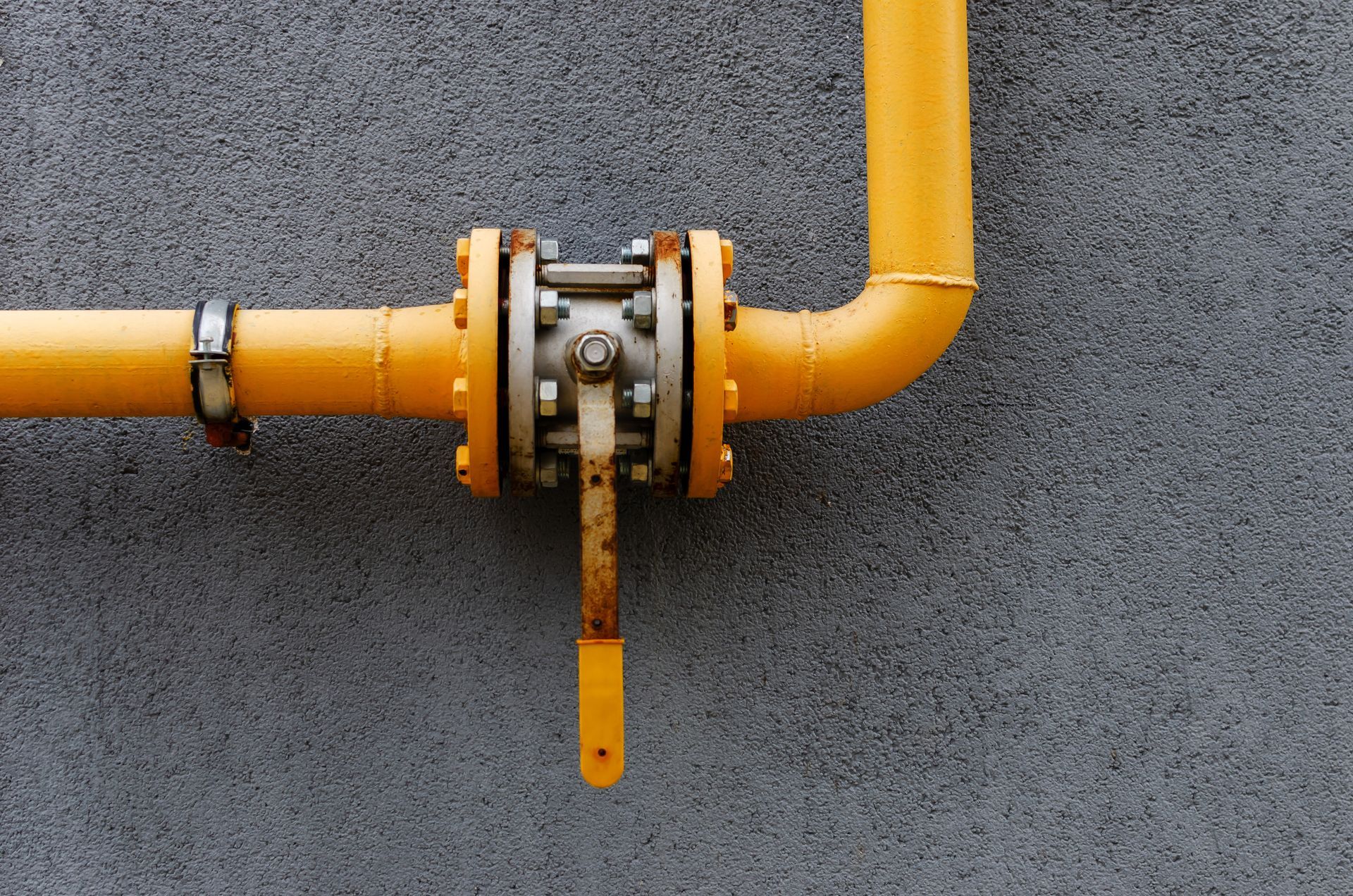 A yellow pipe mounted on a gray textured wall features a large, flanged ball valve with a vertical handle.