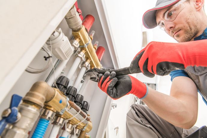 A technician in safety gear uses a wrench to adjust a wall-mounted heating system manifold with copper and white pipes.