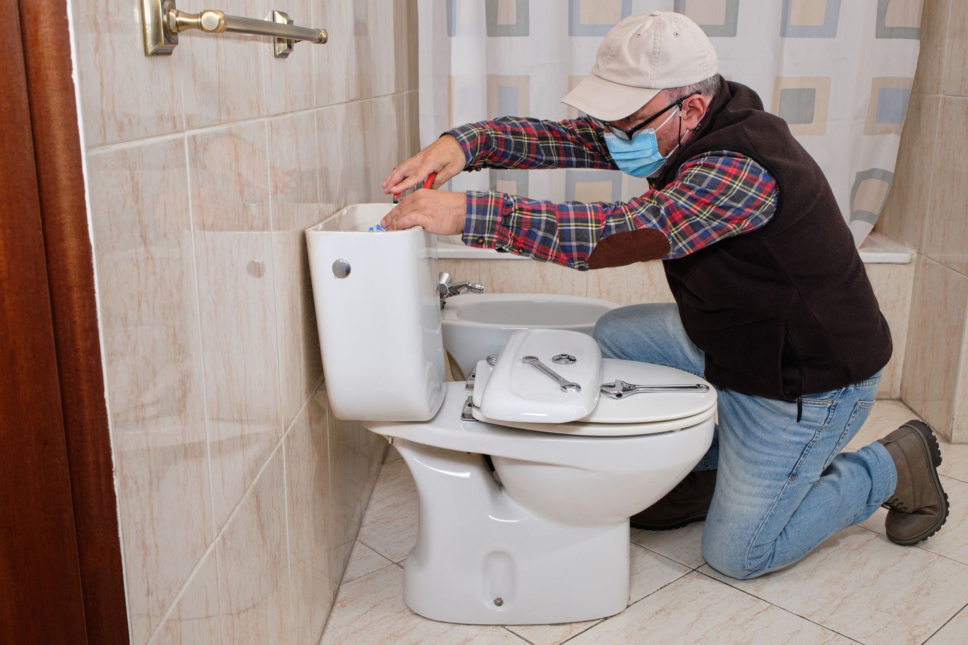 A person wearing a plaid shirt, vest, and face mask kneels in a bathroom to repair an open toilet tank with a wrench.