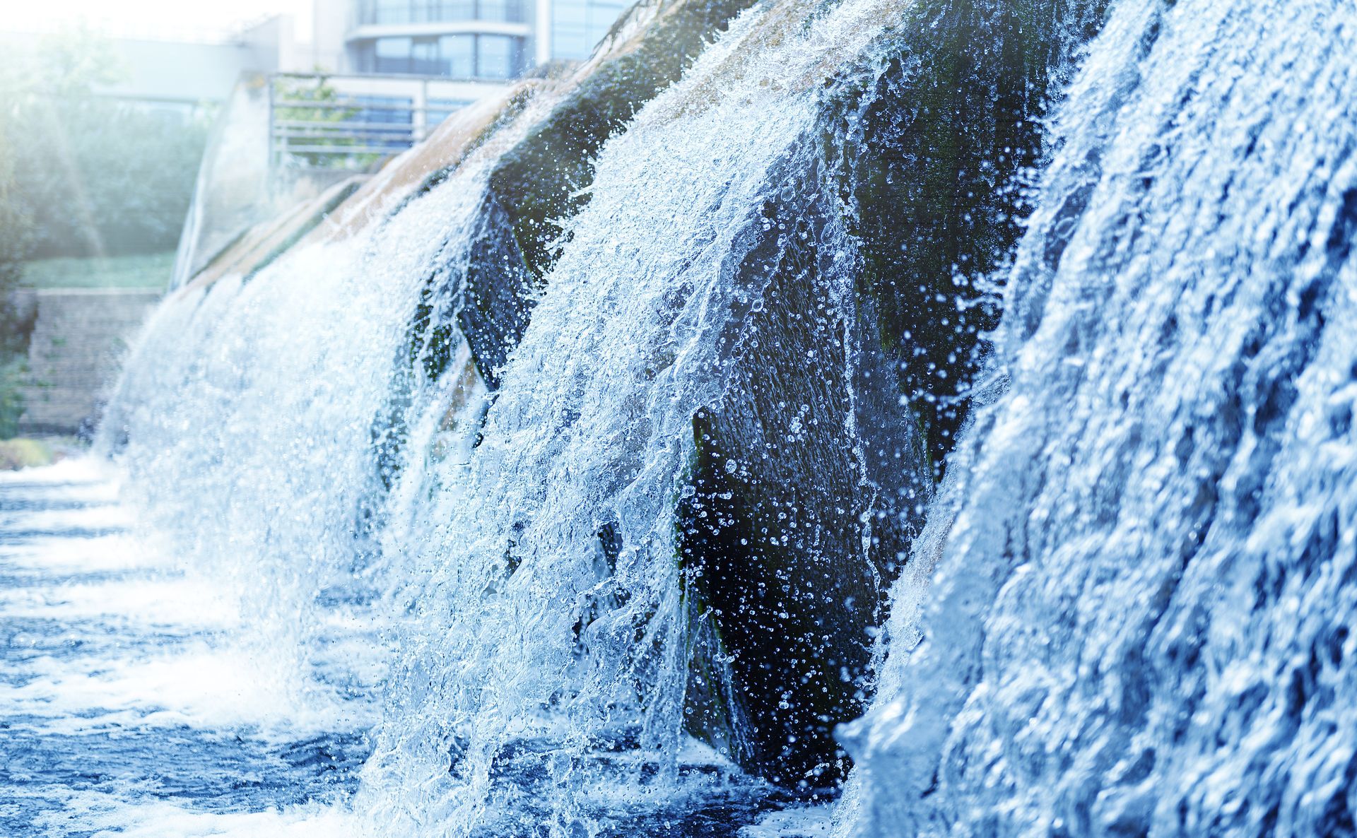 Water flows over a tiered concrete weir or spillway, creating cascading falls in a bright, outdoor setting.