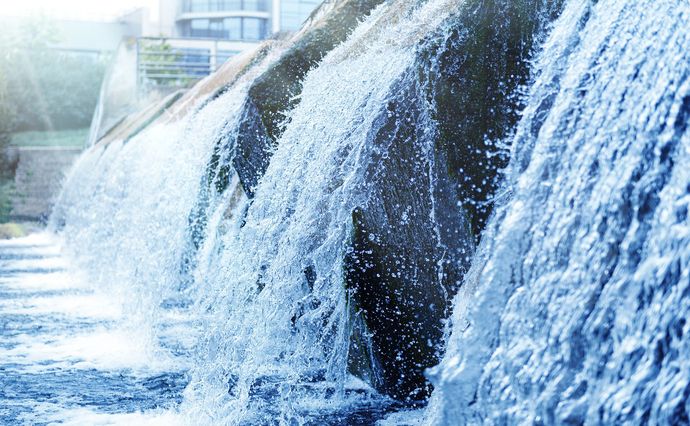 Water flows over a tiered concrete weir or spillway, creating cascading falls in a bright, outdoor setting.