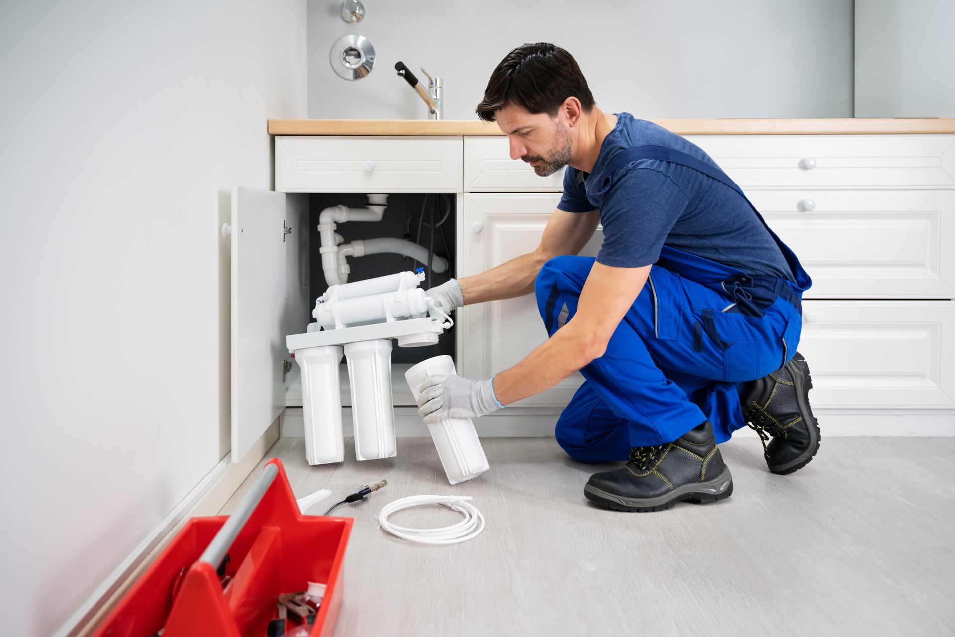 A technician in blue workwear kneeling under a kitchen sink to install a water filtration system.