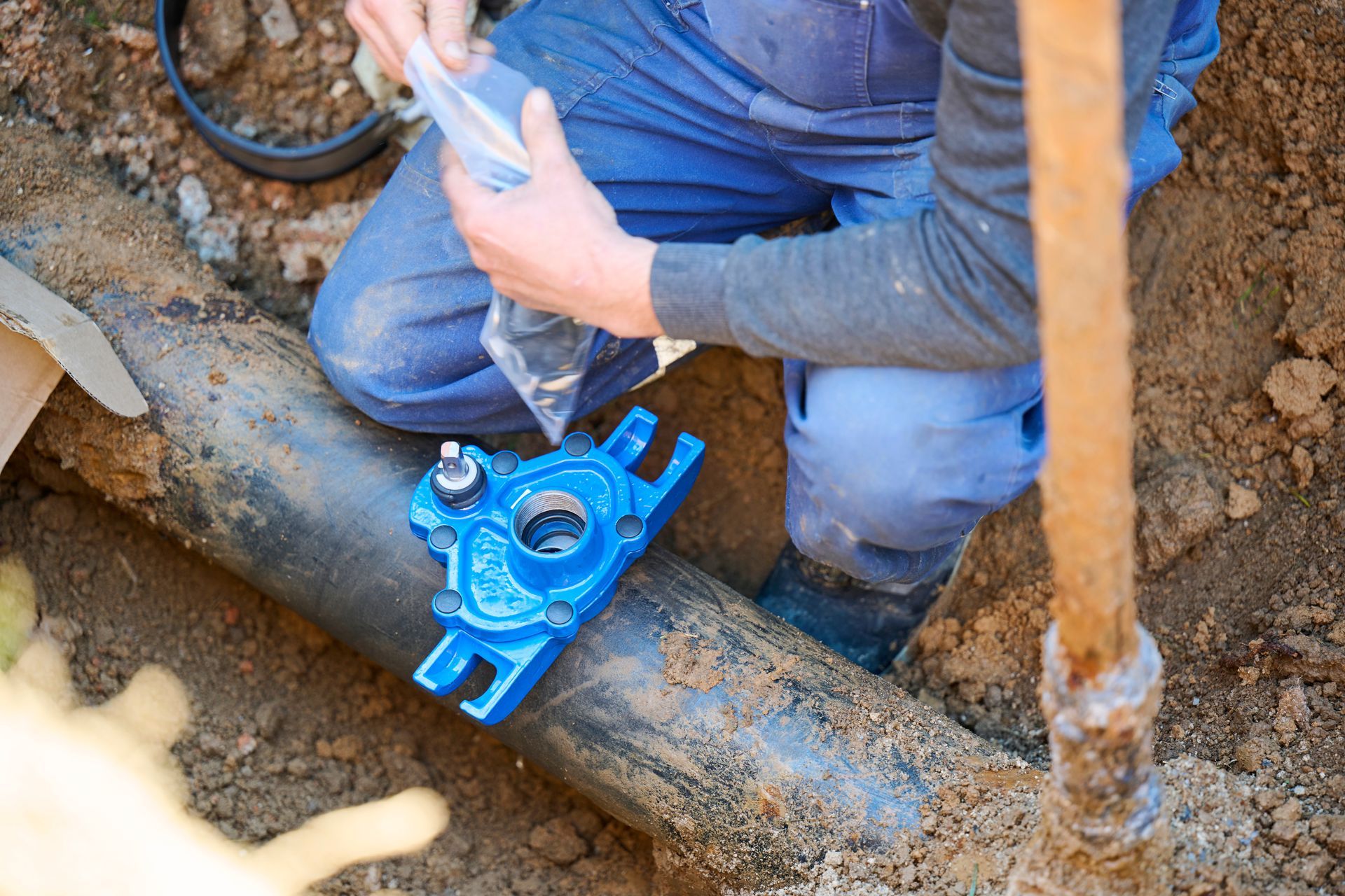 A person in work clothes kneeling in a trench while installing a blue service saddle onto a black pipe.