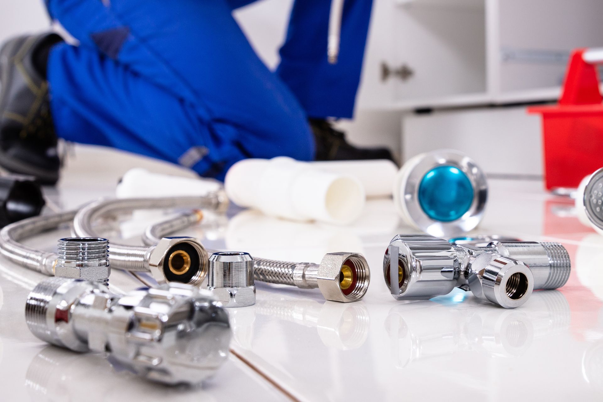 A technician in blue coveralls works near plumbing supplies, including metal pipes and valves, on a white tiled floor.
