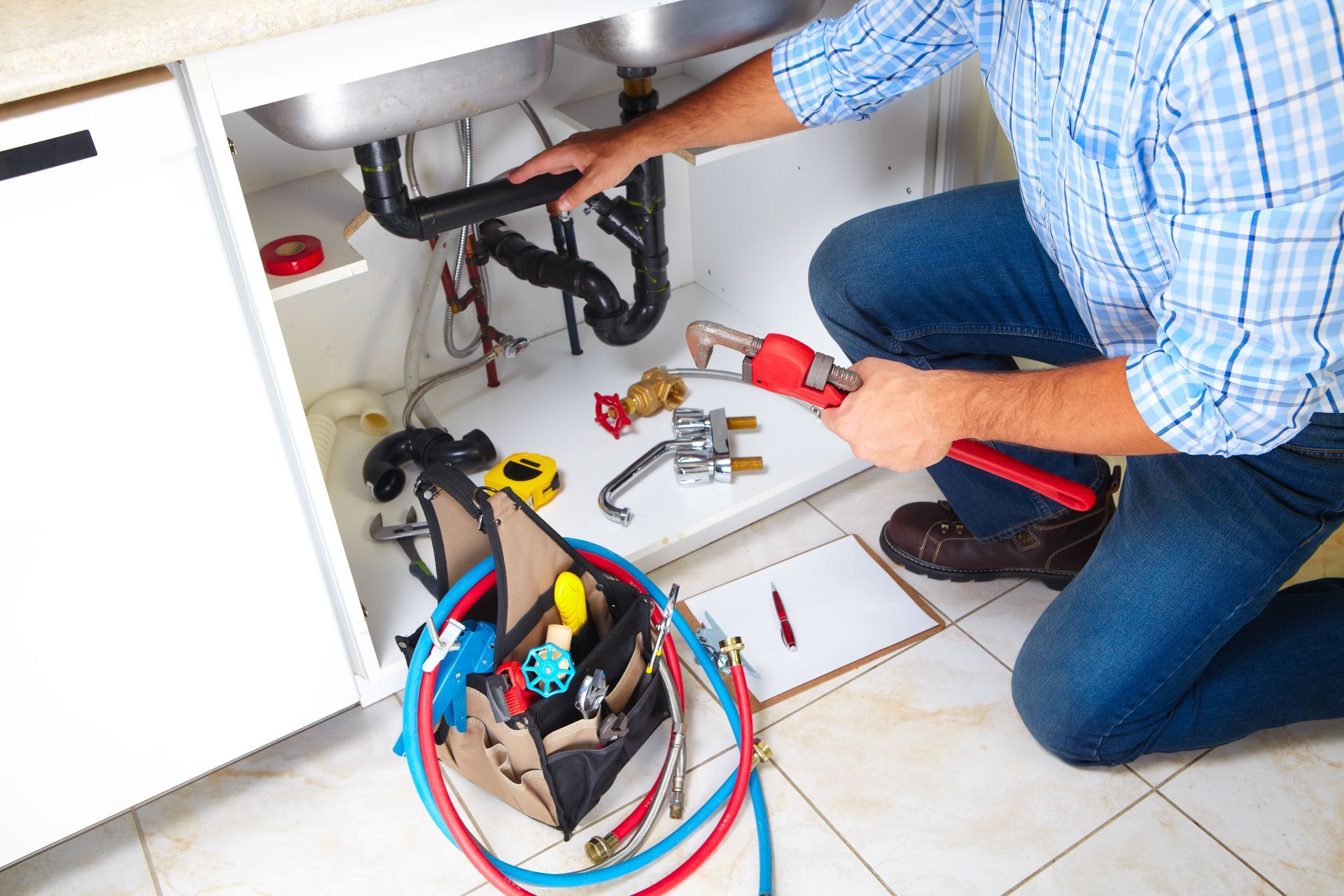 A person in a plaid shirt kneels under a kitchen sink with plumbing tools, a toolbox, and a notepad on the floor.