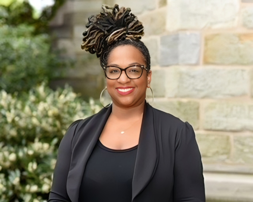 A woman wearing glasses and a black jacket is standing in front of a brick wall.