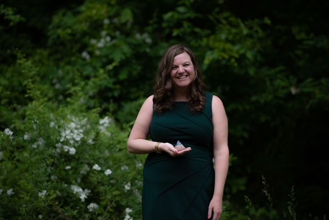 A woman is standing in the woods with her hand on her chest.
