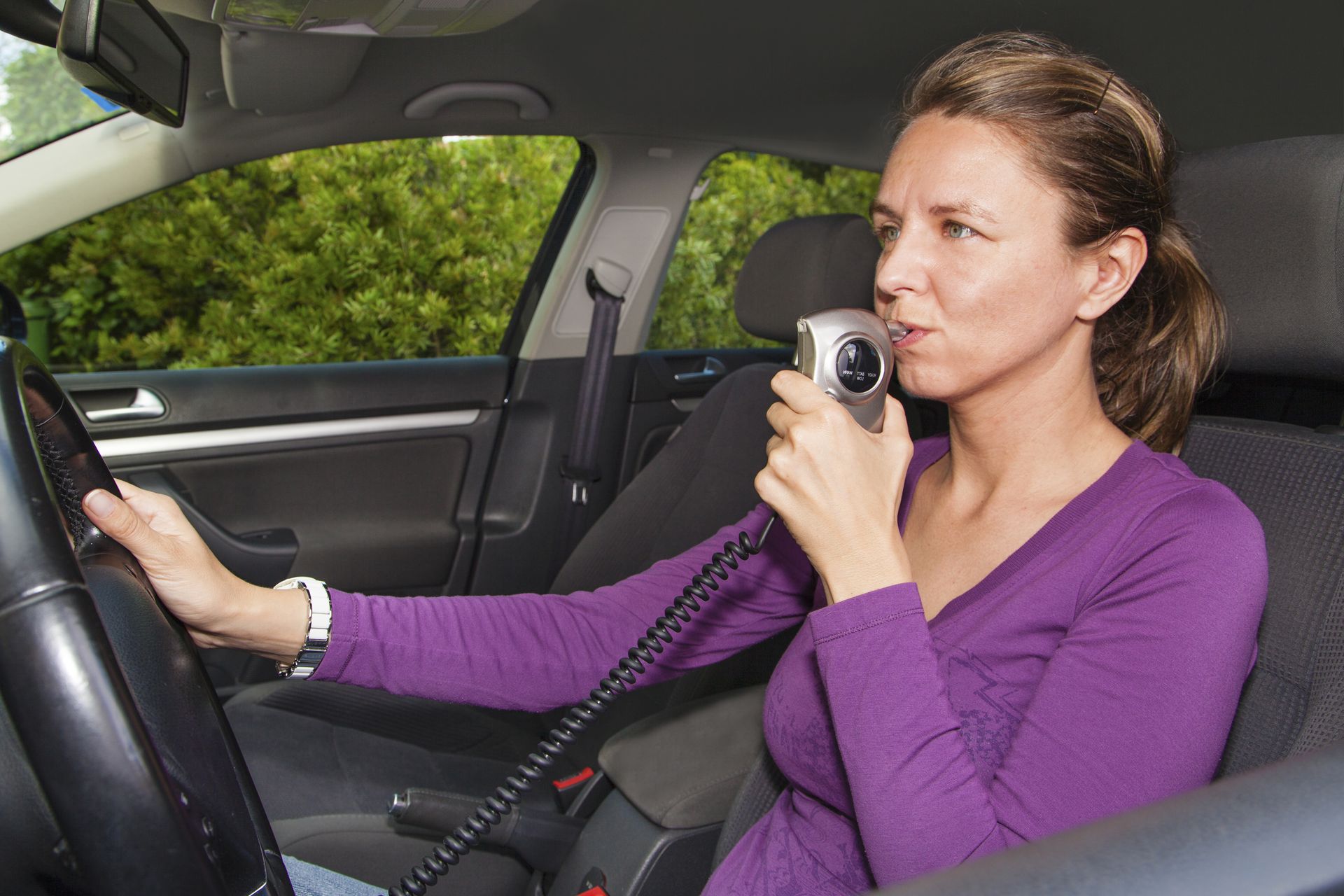 A woman in a purple shirt sitting in her car blowing into an interlock device