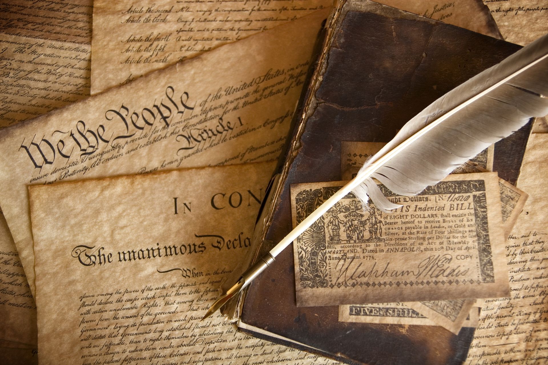 An image of a writing quill on top of a book which is laying on the United States Constitution