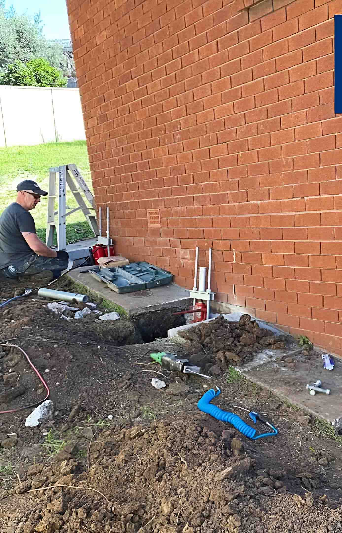 A Man Is Kneeling In The Dirt Next To A Brick Wall — Affordable Underpinning Solutions In Shell Cove, NSW