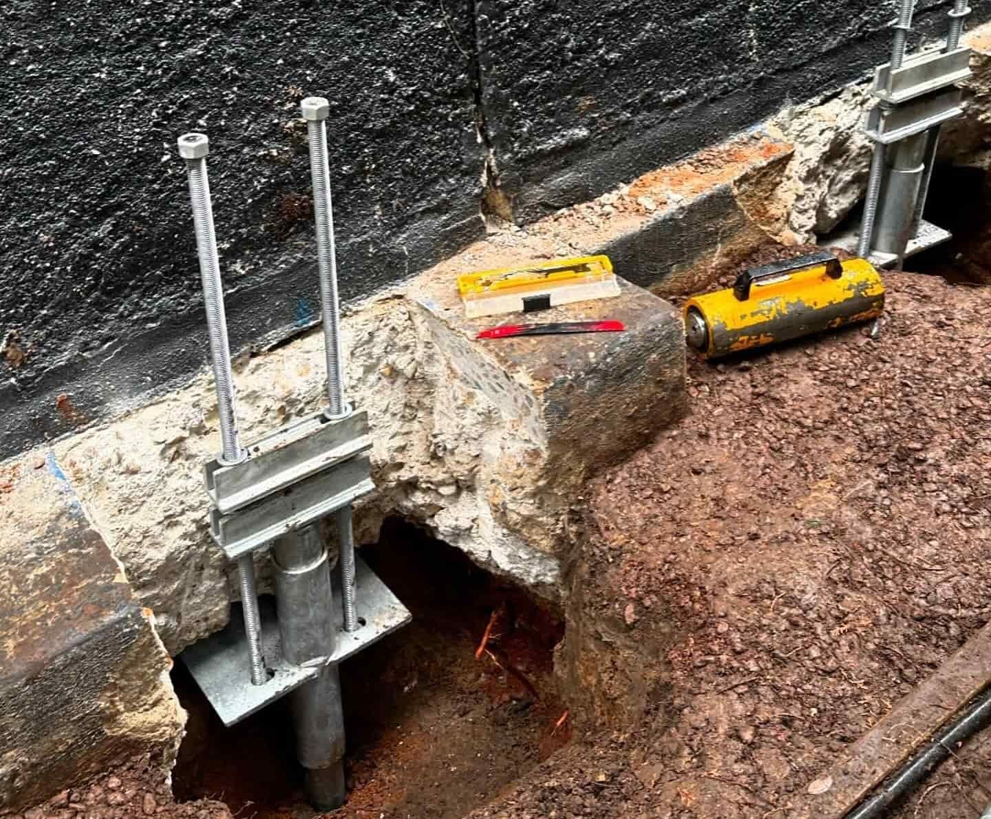 A Metal Post Is Sitting In Dirt Beside a Brick Wall — Affordable Underpinning Solutions In Shell Cove, NSW