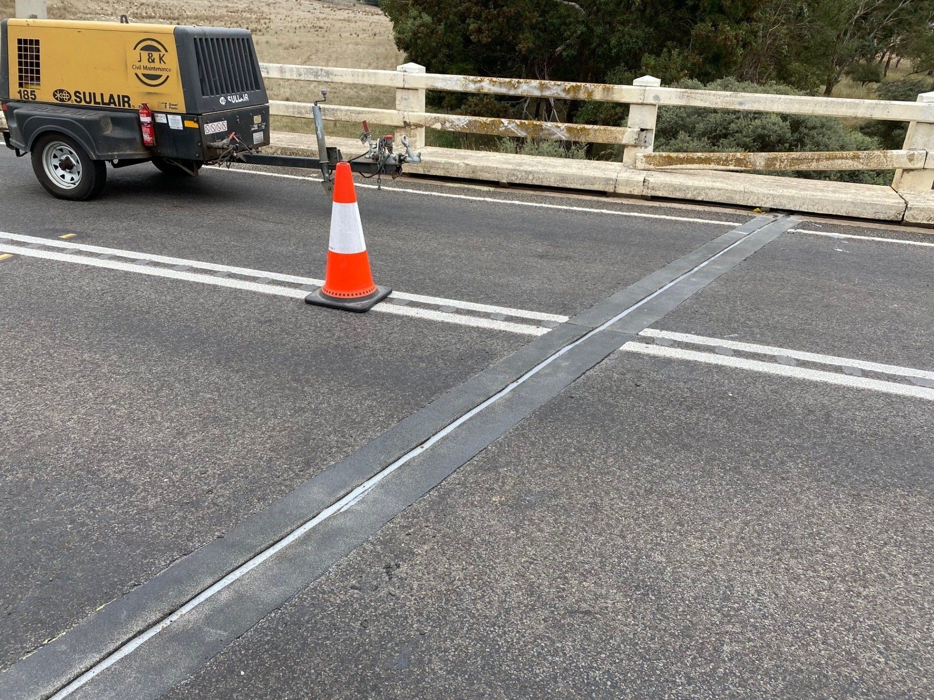 Orange traffic cone on a road with a parked yellow utility trailer near a bridge railing.