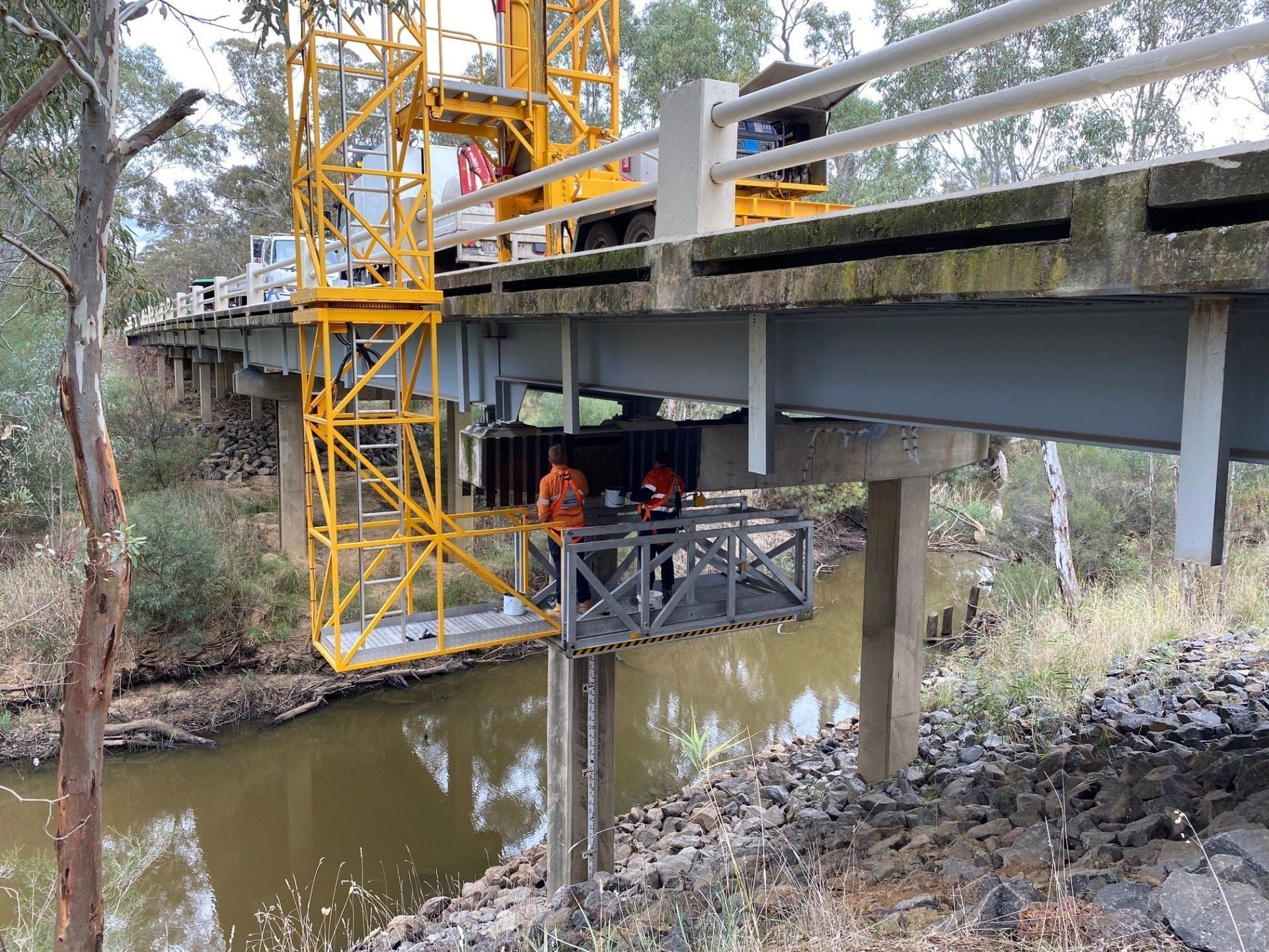 Workers on a bridge construction platform above a muddy creek, with yellow crane scaffolding nearby.