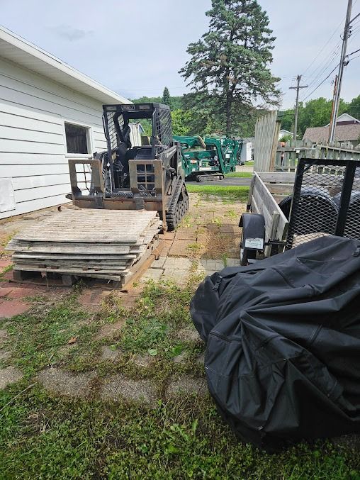 Skid steer loader next to building and trailer; wood pallets and black tarp visible.