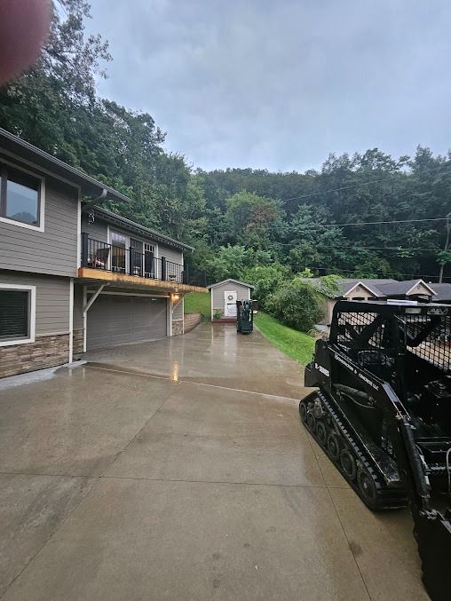 Gray house with a garage and deck, a small shed, and a skid steer on a wet driveway surrounded by trees.