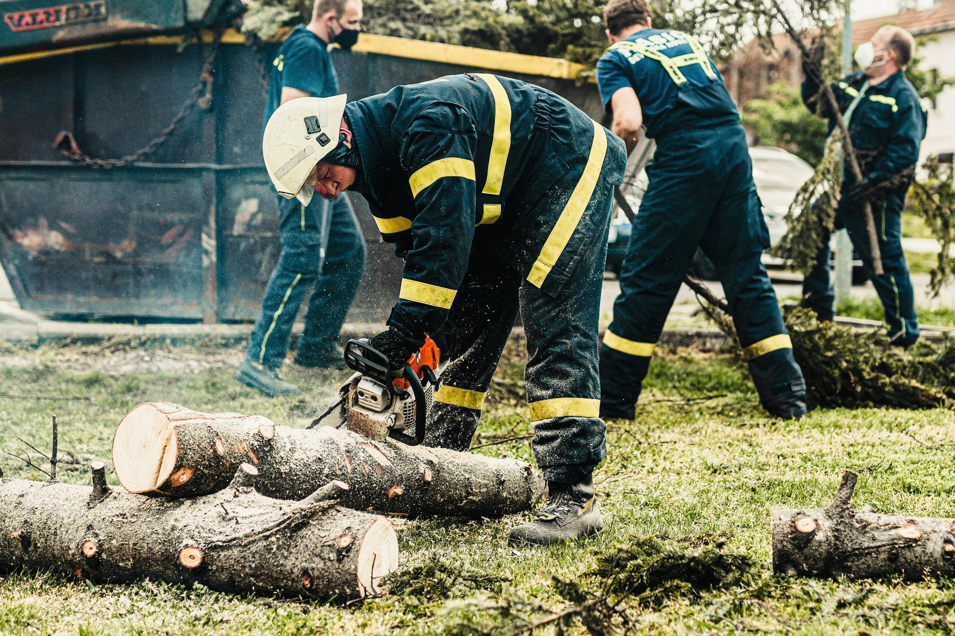 Firefighters using a chainsaw to cut fallen tree trunks in a grassy area.