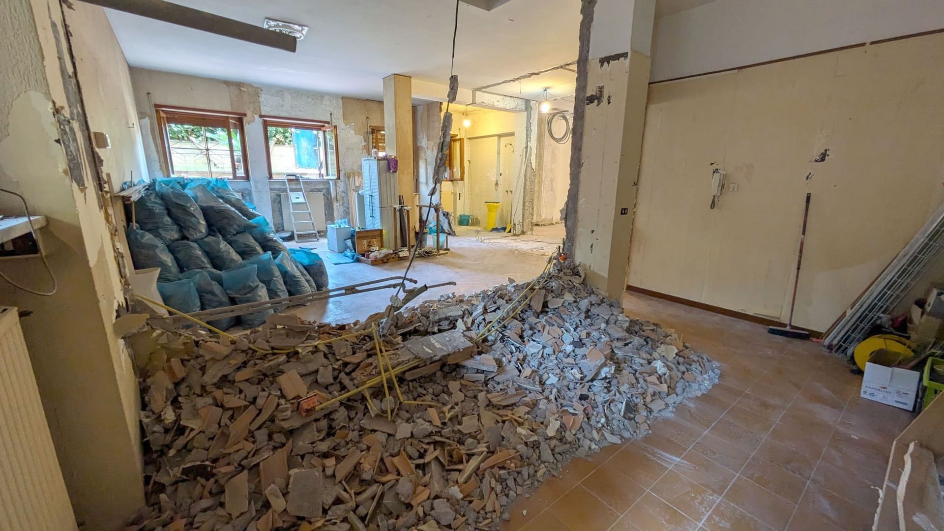 Stanza in fase di ristrutturazione con macerie ammucchiate sul pavimento, pareti a vista e un divano blu in fondo.