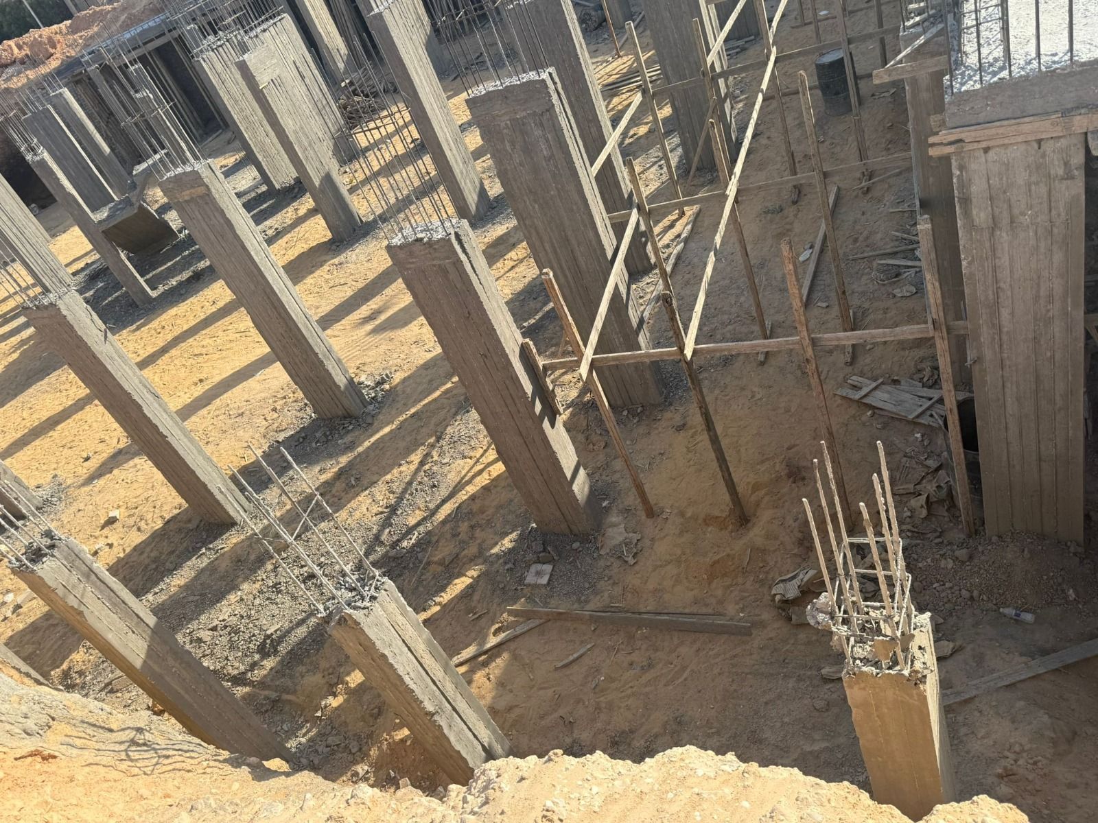 Cantiere edile con colonne in cemento armato e fondamenta in tondini di ferro in uno scavo polveroso.