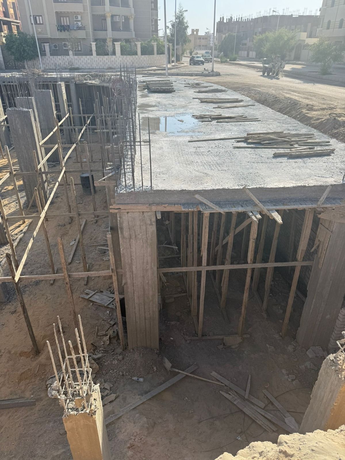 Cantiere edile con fondamenta in cemento, casseforme in legno e colonne di armatura a vista sotto la luce diretta del sole.