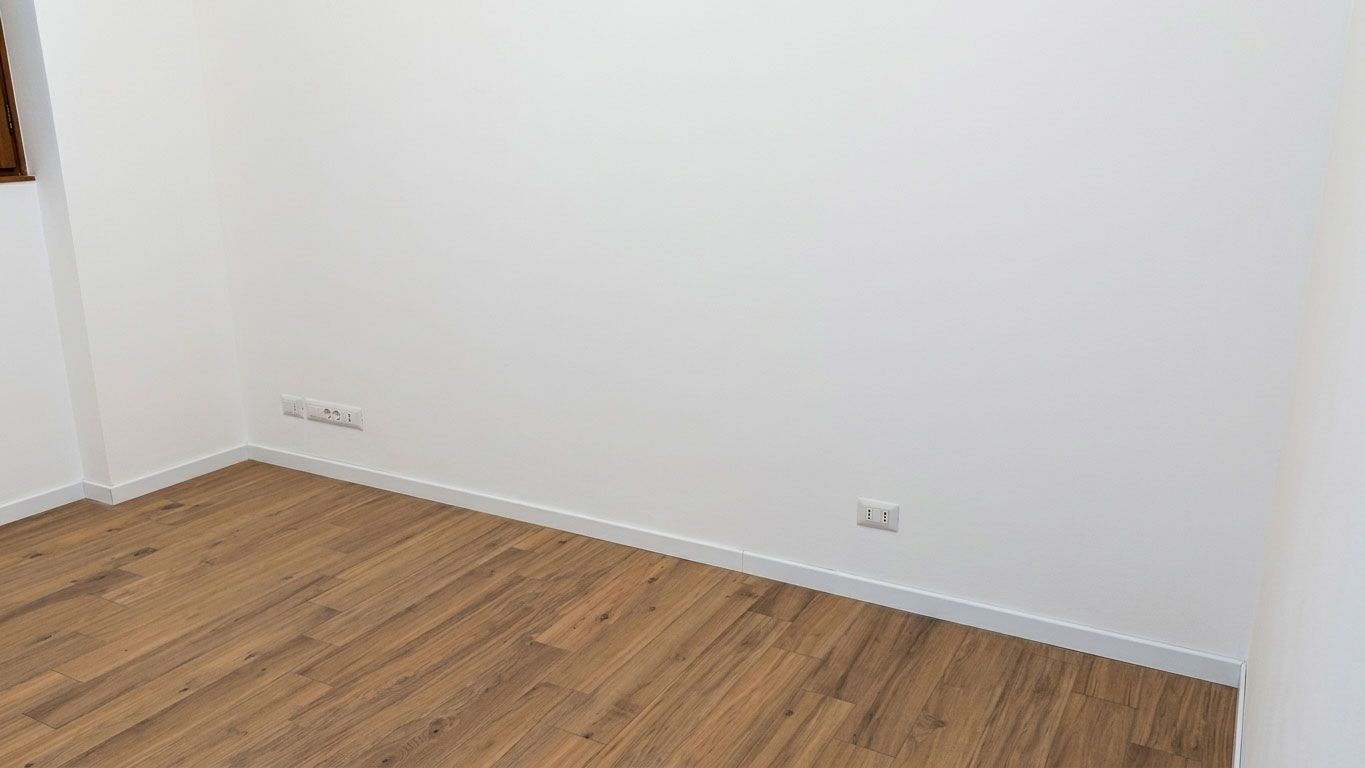 Stanza vuota con pareti bianche, pavimento in legno e prese elettriche lungo il battiscopa.