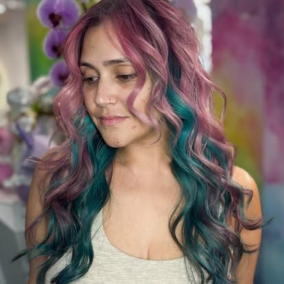 A woman with long pink and blue hair is wearing a white tank top.