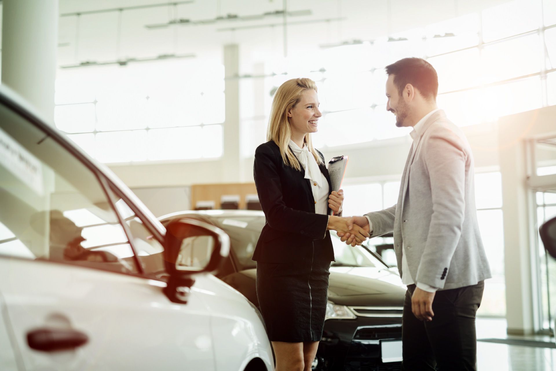 Mujer estrechando la mano a un hombre en un concesionario de automóviles, sonriendo, con coches de fondo.