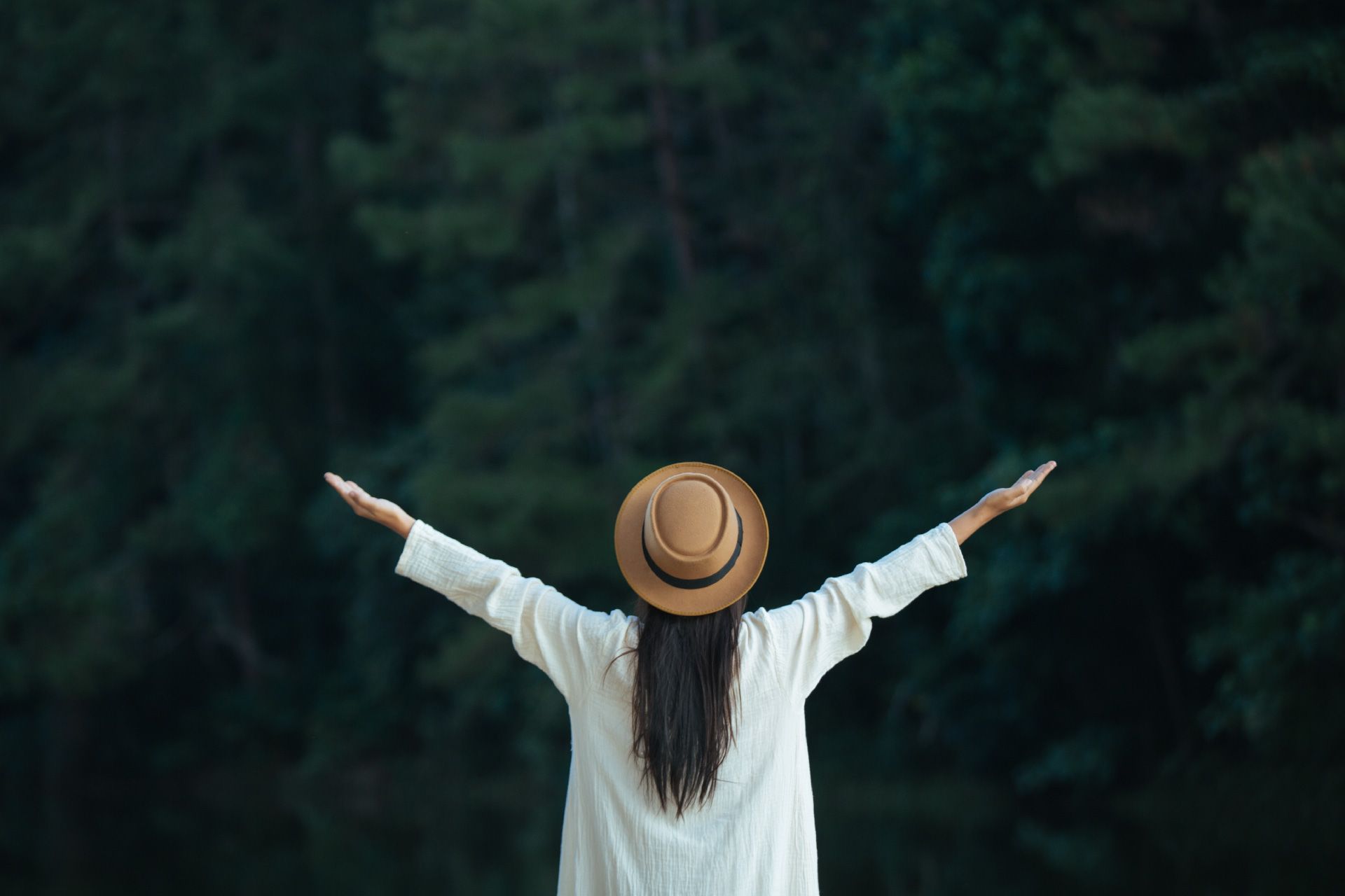 Mujer con sombrero y los brazos levantados, de pie al aire libre, fondo de bosque.