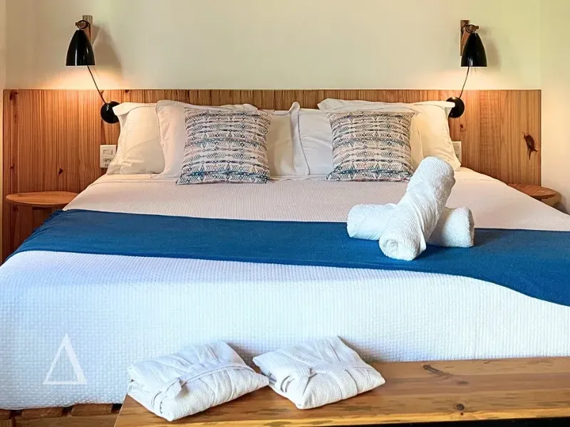 Quarto aconchegante na Quattuor Cabanas com cama king size, enxoval premium branco e azul, cabeceira de madeira e luminárias modernas em São Miguel do Gostoso.