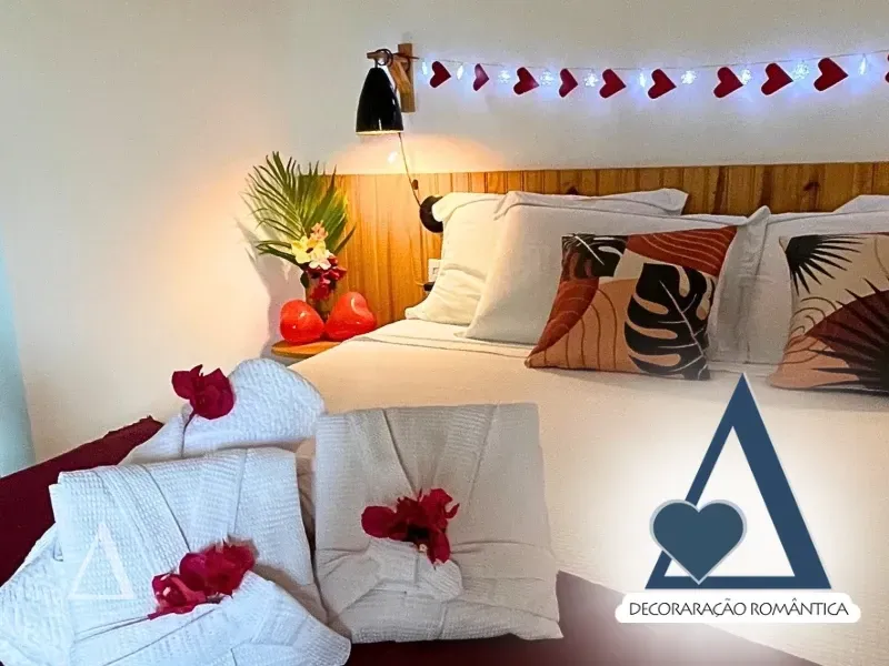 Cama de casal com enxoval branco, pétalas de flores, roupões dobrados e iluminação decorativa de corações na Quattuor Cabanas em São Miguel do Gostoso.