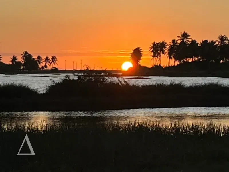 Vista do pôr do sol na Lagoa do Cardeiro a partir da Ponta do Santo Cristo em São Miguel do Gostoso, com silhuetas de coqueiros e reflexo do sol na água.