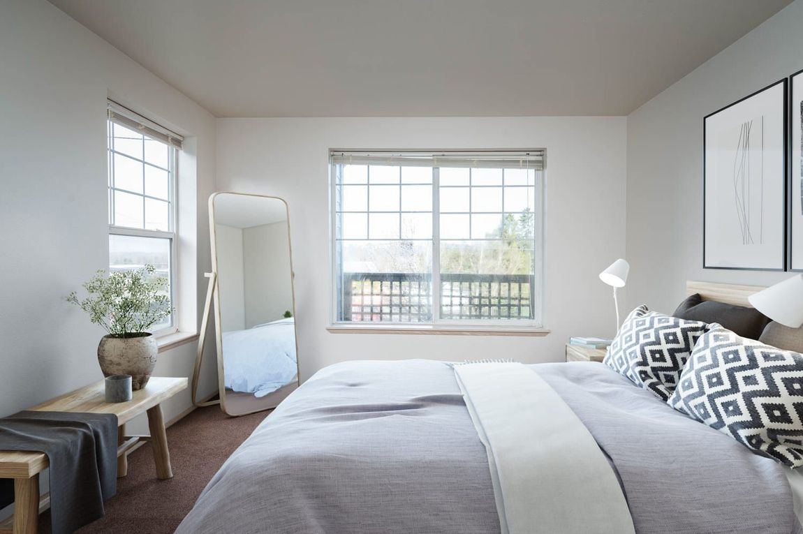 Bedroom with bed, mirror, two windows, art, bench, and neutral color scheme.