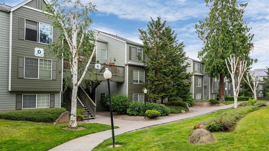 Apartment buildings with green siding, surrounded by trees, grass, and a walking path.