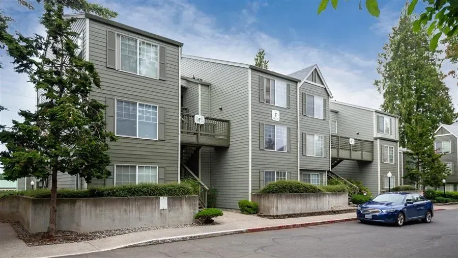 Gray apartment complex with balconies, shrubs, and a blue car parked outside.