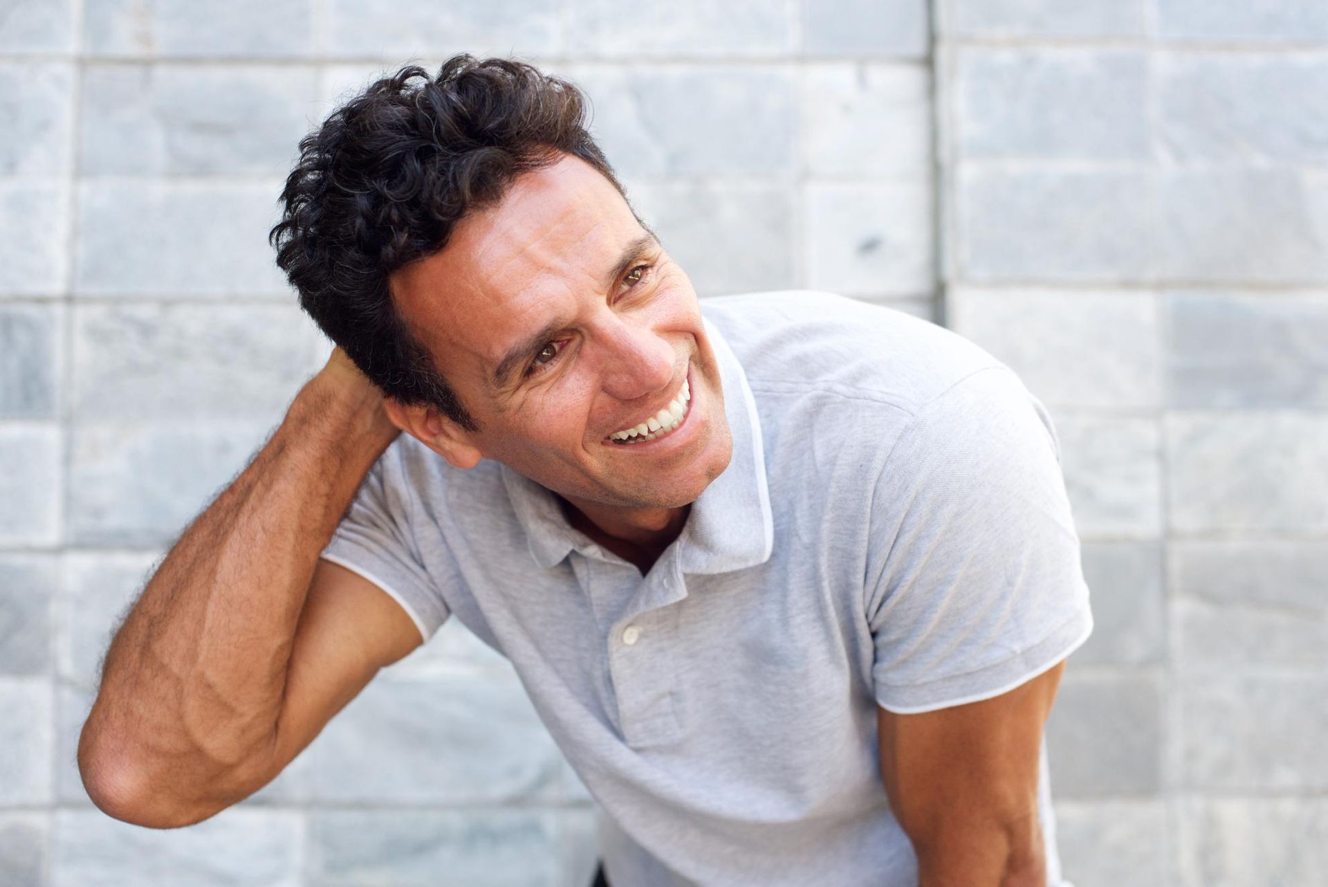 A man in a grey shirt is smiling with his hand on his head.